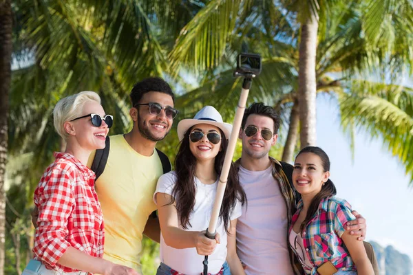 Les gens prennent Selfie groupe avec caméra d'action sur bâton tout en marchant dans Palm Tree Park sur la plage, heureux sourire mélange Race Friends — Photo