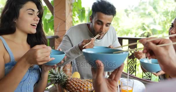 La gente se alimenta mutuamente con deliciosos fideos comida asiática, grupo de amigos se sientan en la mesa en la terraza con vistas al bosque tropical — Vídeo de stock