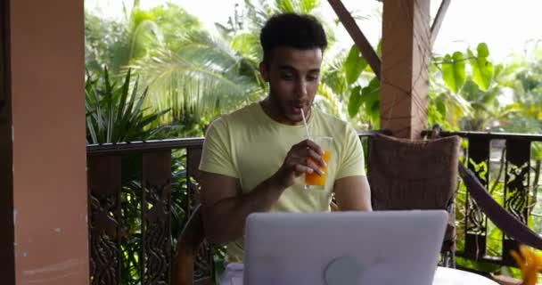 Jovem usa computador portátil no terraço bebendo suco latino homem digitando teclado durante o café da manhã ao ar livre — Vídeo de Stock