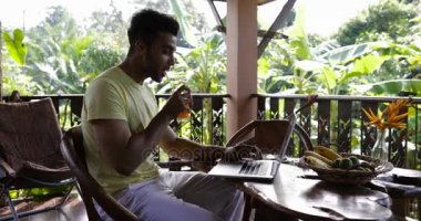 Genç adam Teras Mix yarış adam içecek suyu çalışırken tablo açık havada oturup kahvaltı dizüstü bilgisayarda yazarak