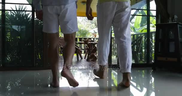 Dos hombres caminando en la terraza sentados en la mesa hablando beber jugo de naranja, mezcla de raza gay pareja por la mañana en villa en el bosque tropical — Vídeos de Stock