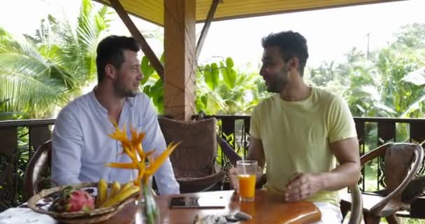 Dos hombres sentados en la terraza de verano hablando beber zumo de naranja, mezcla de raza gay pareja en la mañana en Villa en el bosque tropical — Vídeo de stock