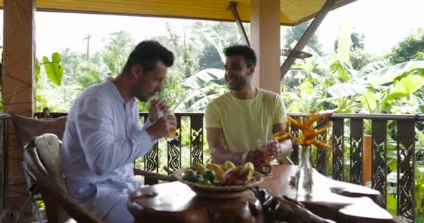 Zwei Männer sitzen auf Sommerterrasse reden halten digitalen Tablet-Computer, mischen Rennen schwules Paar in der Früh auf Villa im tropischen Wald — Stockvideo
