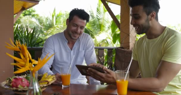 Due uomini che utilizzano computer tablet seduti sulla terrazza estiva a parlare, giovani ragazzi casual touch screen surf web insieme — Video Stock