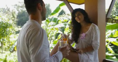 Erkek ve kadın güzel tropik orman villada sabah Toasting Hotel, mutlu balkonda konuşurken genç çift İçecek suyu