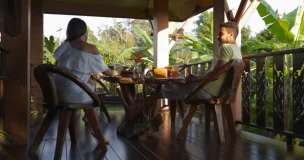 Pareja joven sentada en la terraza disfrutando de la puesta del sol Vista del bosque tropical Hablando y golpeando vasos de jugo, hombre feliz y mujer — Vídeos de Stock