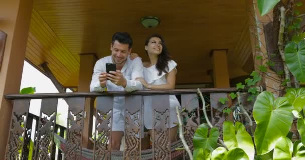 Νεαρό ζευγάρι στο μπαλκόνι, κοιτώντας ψηλά μιλάμε άνδρας και η γυναίκα αγκαλιάζει κρατήστε χρήση κυττάρων έξυπνο τηλέφωνο ευτυχισμένη χαμογελώντας — Αρχείο Βίντεο