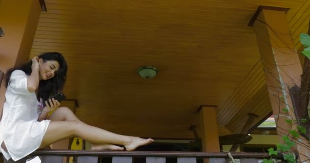 Junge Frau benutzt Handy auf Balkon sitzend glücklich lächelnd, schönes Mädchen chattet online im Freien — Stockvideo