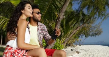 İki avuç içi hücre akıllı telefon kullanarak, mutlu gülümseyen adam ve kadın söz altında sahilde kucaklayan oturmak