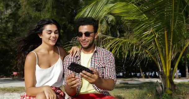 Pareja usando celular teléfono inteligente tomar foto selfie al aire libre bajo las palmeras en la playa, feliz hombre y mujer sonriente — Vídeos de Stock