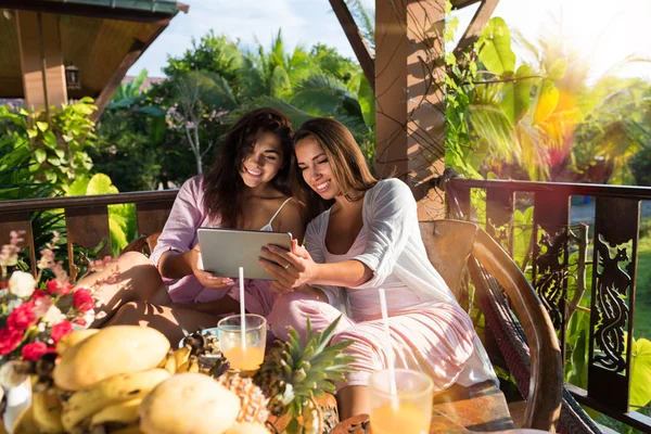 Dos mujeres jóvenes usan tableta digital mientras desayunan en la terraza en el jardín tropical Hermosas chicas sostienen la PC móvil inalámbrica sentadas juntas en la mesa Ver fotos —  Fotos de Stock