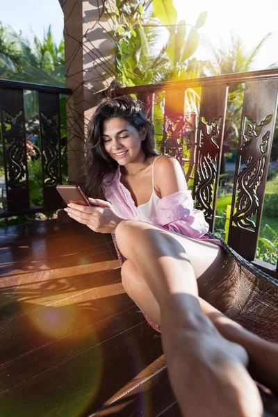 Молодая красивая женщина в гамаке на летней террасе использовать мобильный смартфон счастливая улыбающаяся привлекательная девушка расслабляясь — стоковое фото