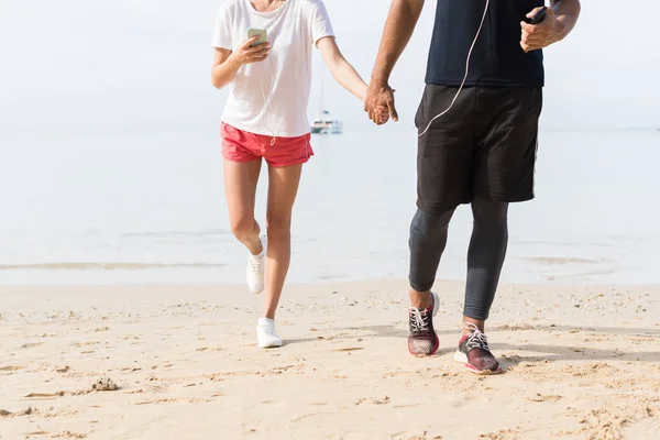 Αγνώριστος ζευγάρι των δρομέων, κρατώντας τα χέρια χρήση κυττάρων έξυπνα τηλέφωνα άνδρας και γυναίκα αθλητισμού δρομείς στην παραλία εργάζονται έξω μίγμα αγώνα Jogger ανδρικά και γυναικεία Fitness — Φωτογραφία Αρχείου