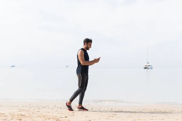 Tiro de comprimento total do corredor masculino usando o telefone inteligente celular na praia enquanto jogging no homem do litoral Jovem esporte latino — Fotografia de Stock