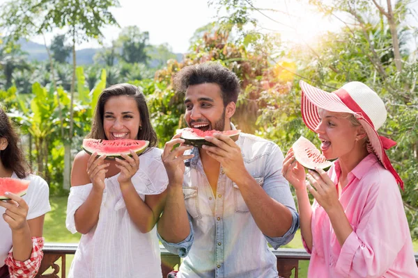 Весела група друзів їдять кавун разом на відкритому повітрі на літній терасі з тропічним лісом тло мікс гонки люди розважаються — стокове фото
