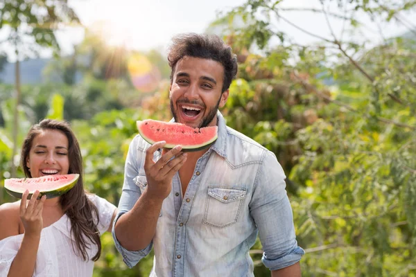 Felice uomo e donna mangiare anguria insieme oltre bella tropicale foresta paesaggio allegro coppia ridere tenendo fetta di anguria — Foto Stock