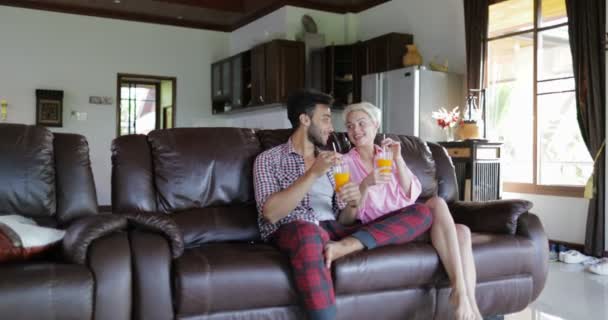 Пара сидящих на тренере в гостиной пить апельсиновый сок, молодой человек и женщина по утрам говорить современные квартиры студии внутренних — стоковое видео