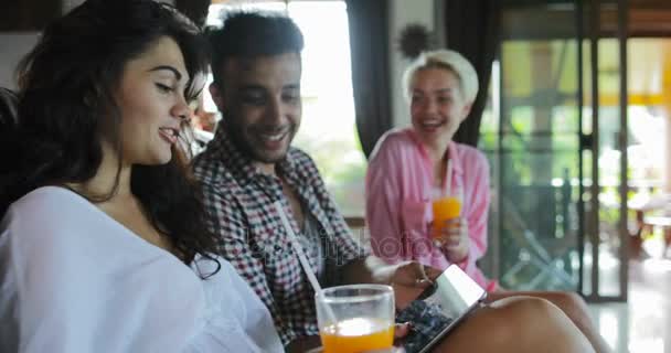 La gente usa la computadora de la tableta en el entrenador que mira la televisión en la sala de estar bebe el jugo, el hombre joven y el grupo de la mujer en el interior moderno del apartamento que habla de la mañana — Vídeos de Stock