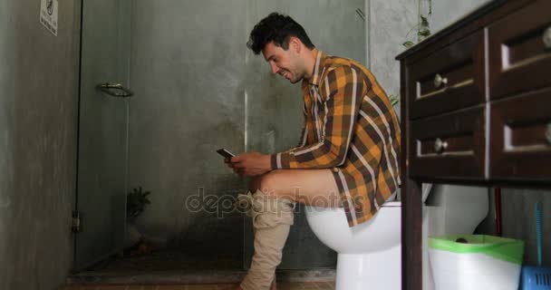 Ο άνθρωπος που κάθεται στην τουαλέτα χρησιμοποιούν κινητό έξυπνο τηλέφωνο νεαρός άντρας κουβέντα Online — Αρχείο Βίντεο