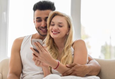 Genç güzel çift pencere, yakınındaki Koç kullanarak hücre akıllı telefon mutlu gülümseme İspanyol kadın erkek oturmak