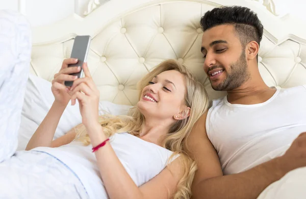 Junges Paar liegt im Bett, glückliches Lächeln spanischer Mann und Frau mit Handy — Stockfoto