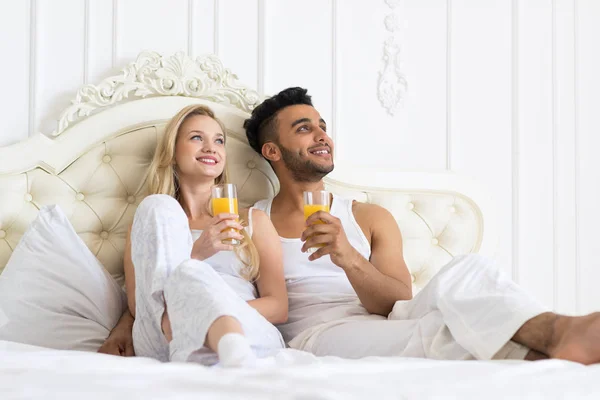 Mladý pár pít Orange Juice sedí postel, šťastný úsměv mladé hispánské muž a žena ve snu Hledat kopie prostor — Stock fotografie