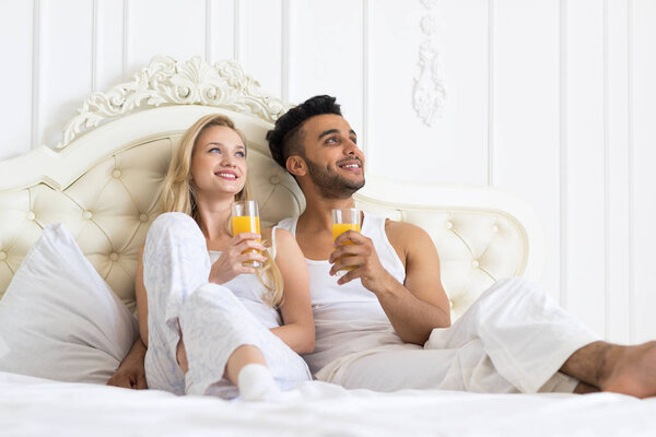 Young Couple Drink Orange Juice Sitting Bed, Happy Smile Young Hispanic Man And Woman dream Look Up Copy Space