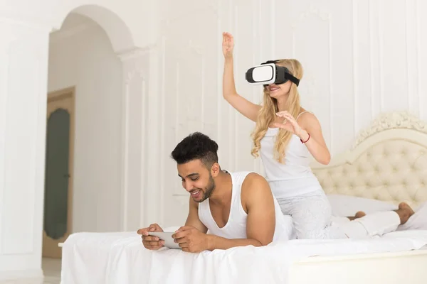 Νεαρό ζευγάρι στο κρεβάτι, Ισπανόφωνος άνθρωπος χρησιμοποιώντας κινητό έξυπνο τηλέφωνο γυναίκα φορούν ακουστικά ψηφιακής γυαλιά εικονικής πραγματικότητας — Φωτογραφία Αρχείου