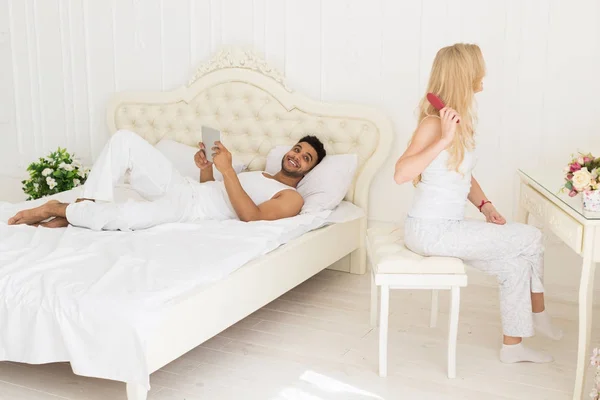 Giovane coppia in camera da letto, ispanico uomo utilizzando tablet computer sdraiato letto e donna seduta spazzolatura capelli — Foto Stock