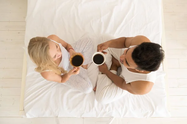 年轻夫妇喝咖啡坐在床上, 快乐的微笑拉美裔男人和女人的顶角视图 — 图库照片