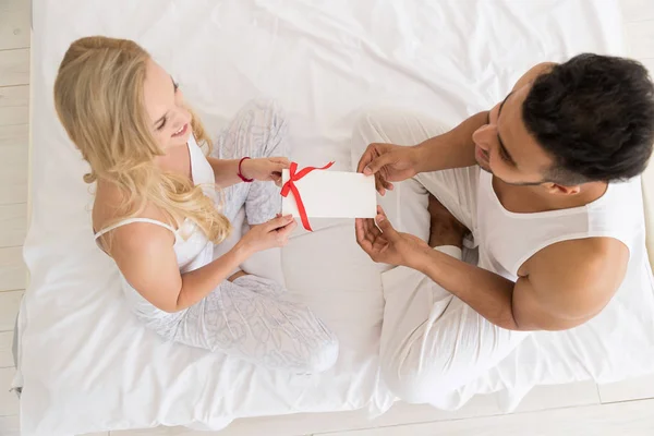 Joven pareja feliz sentada en la cama, el hombre hispano le da a la mujer un sobre sorpresa con cinta, celebración del aniversario —  Fotos de Stock