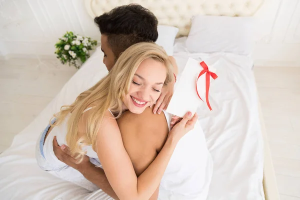 年轻幸福的情侣抱在床上, 拉美裔妇女举行礼物信封带丝带, 周年庆典 — 图库照片
