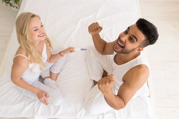 Joven pareja sentada en la cama, feliz sonrisa mujer espectáculo excitado gritando sorprendido hombre positivo embarazo prueba — Foto de Stock