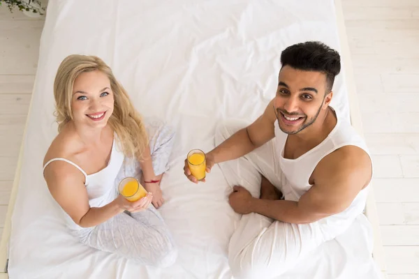 Mladý pár pít pomerančový džus sedí v posteli, šťastný úsměv hispánský muž a žena vrcholový úhel pohledu — Stock fotografie