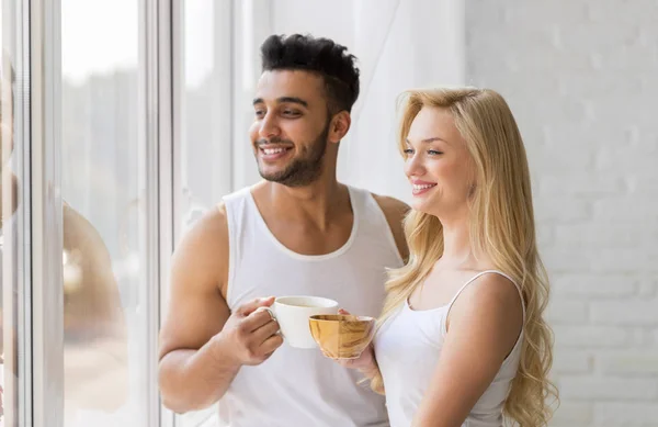 Красива молода пара стенд поблизу великі вікна, пити вранці кавові чашки, щасливою усмішкою іспаномовні чоловік жінка — стокове фото