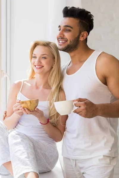 Mladý nádherný pár stát poblíž velké okno, pijte ráno šálek kávy, šťastný úsměv hispánské muž žena — Stock fotografie