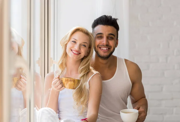 Junges schönes Paar steht am großen Fenster, trinkt Morgenkaffeetasse, fröhliches Lächeln spanischer Mann Frau — Stockfoto