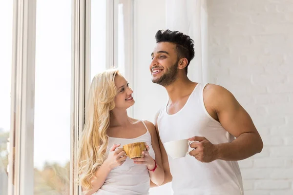 Pareja joven y hermosa se para cerca de una ventana grande, bebe taza de café por la mañana, sonrisa feliz mujer hispana —  Fotos de Stock