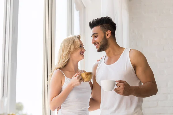 Unga vackra paret stå nära stora fönster, dricker morgon kaffekopp, lyckligt leende spansktalande Man kvinna — Stockfoto