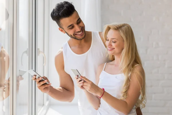Mladý nádherný pár stát poblíž velké okno, pomocí buňky chytrý telefon šťastný úsměv hispánský muž žena — Stock fotografie
