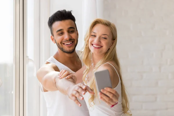 Joven hermosa pareja de pie ventana, tomando foto selfie en el teléfono inteligente celular —  Fotos de Stock