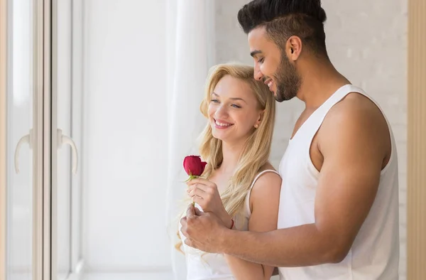 Junges schönes Paar steht am großen Fenster, hispanischer Mann gibt Frau rote Rose — Stockfoto