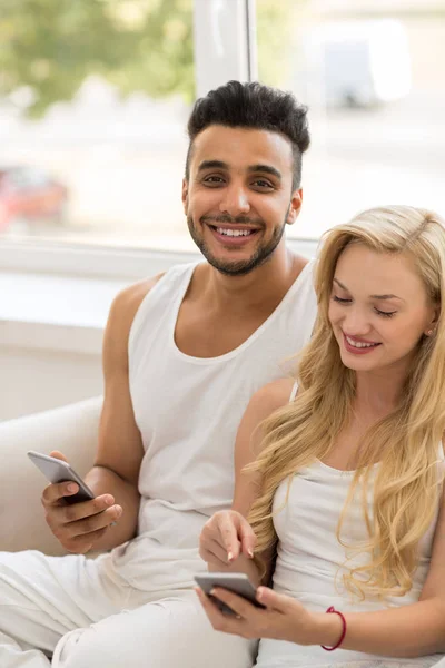 Krásný mladý pár sedí na trenér poblíž okna, pomocí buňky chytrý telefon šťastný úsměv hispánský muž žena — Stock fotografie