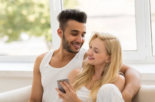 Jonge mooie paar zitten op Coach in de buurt van venster, met behulp van cel slimme telefoon gelukkig glimlach Spaanse Man vrouw — Stockfoto