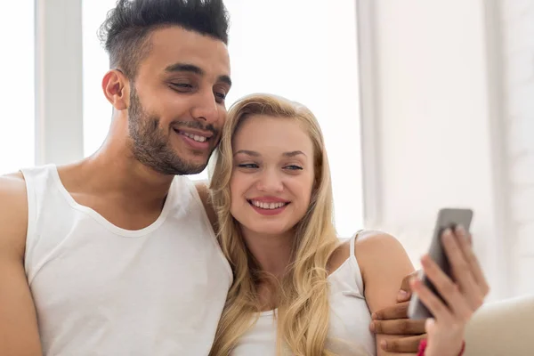 Jonge mooie paar zitten op Coach in de buurt van venster, met behulp van cel slimme telefoon gelukkig glimlach Spaanse Man vrouw — Stockfoto