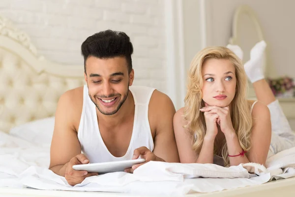 Ungt par ligger i sängen, lyckligt leende spansktalande Man använder tablett dator irriterad uttråkad kvinna — Stockfoto