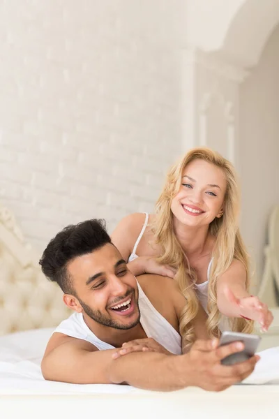 Pareja joven acostada en la cama, hombre hispano sonriente feliz usando el teléfono inteligente celular, mujer tratando de llevarse —  Fotos de Stock