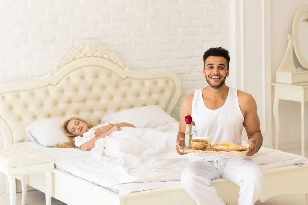 L'homme hispanique apporte le petit déjeuner à la femme endormie dans le plateau du matin avec la fleur de rose rouge, jeune couple — Photo