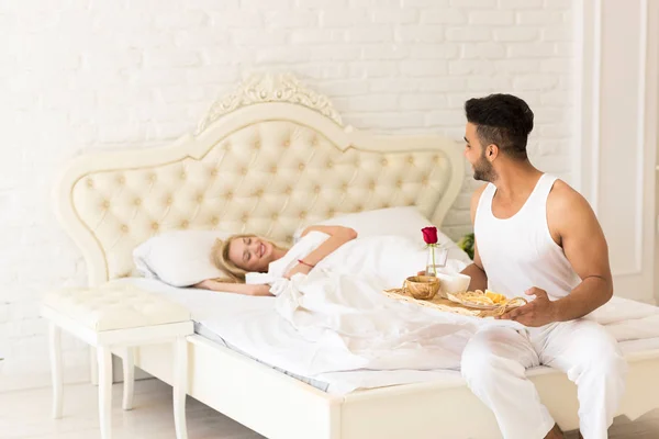 L'homme hispanique apporte le petit déjeuner à la femme endormie dans le plateau du matin avec la fleur de rose rouge, jeune couple — Photo