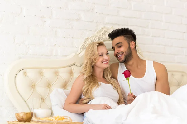 Pareja joven acostada en la cama desayunar por la mañana con flor de rosa roja, feliz sonrisa hispana hombre y mujer —  Fotos de Stock